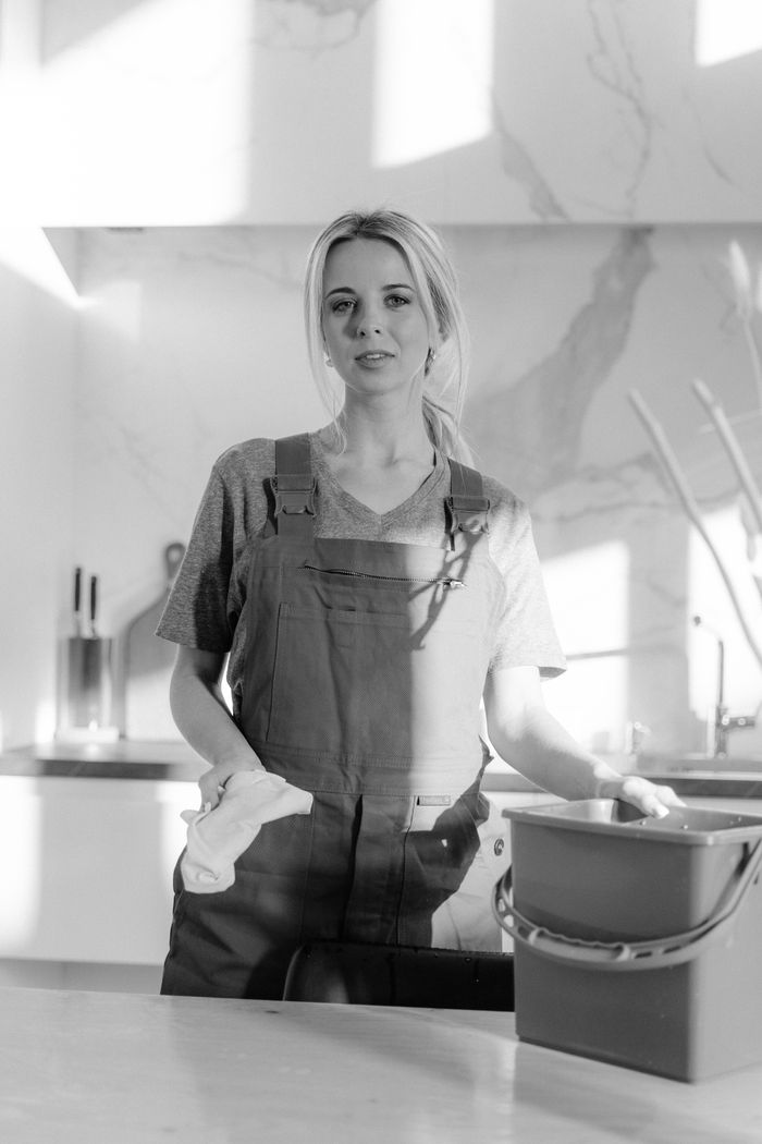 Portrait noir et blanc d'une femme de ménage avec des produits de nettoyage dans une cuisine moderne.